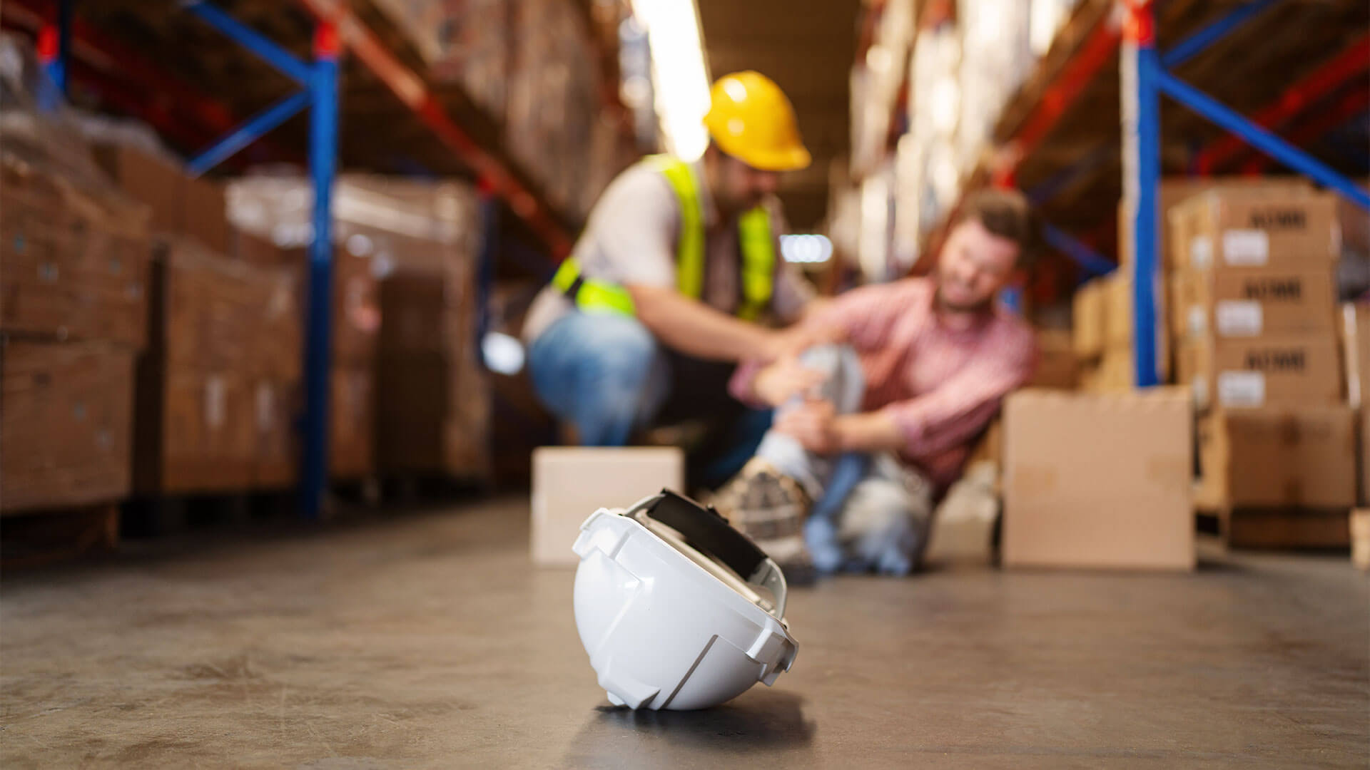 Em primeiro plano um capacete no chão e ao fundo um funcionário de fábrica machucado recebendo ajuda de colega de trabalho